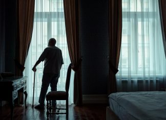 From the Retirement Home, Emailing Love Letters to My Dead Wife Silhouette of an elderly man standing with cane, gazing out of curtain-covered window.