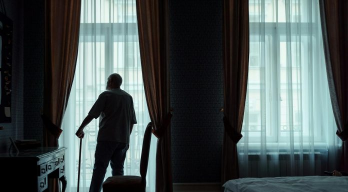 From the Retirement Home, Emailing Love Letters to My Dead Wife Silhouette of an elderly man standing with cane, gazing out of curtain-covered window.