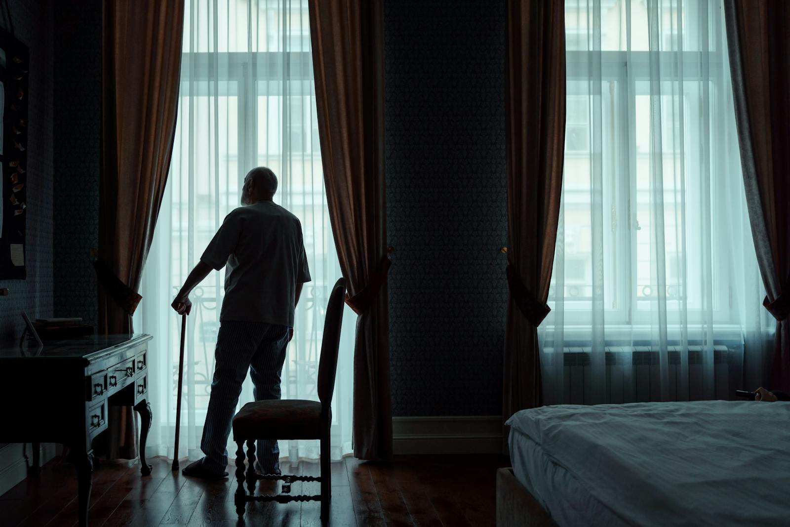 Silhouette of an elderly man standing with cane, gazing out of curtain-covered window.